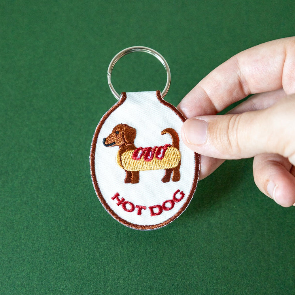 Embroidered keychain with hot dog dachshund design held by a hand against a green background.