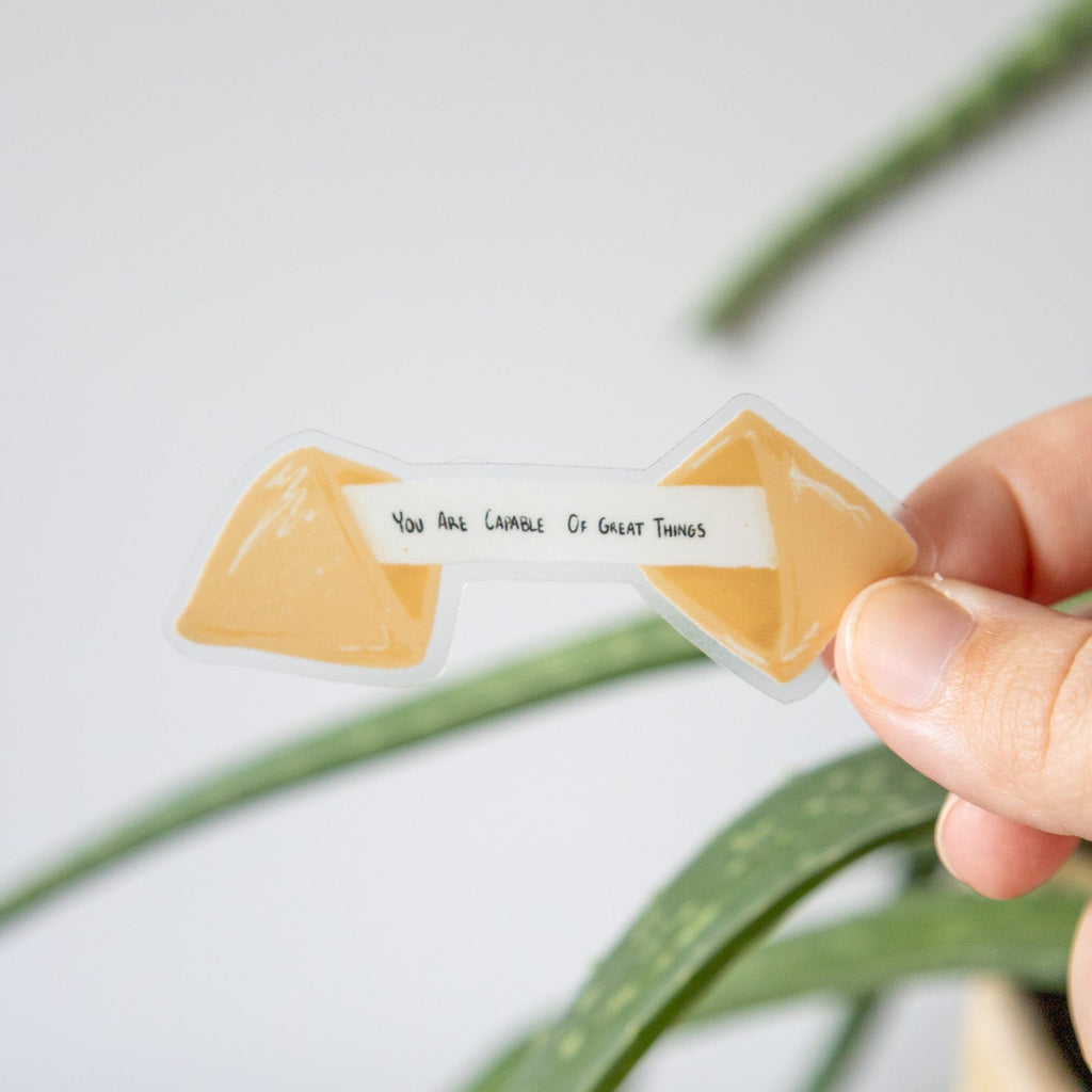 fortune cookie sticker that says "you are capable of great things held by a hand with a plant in the background