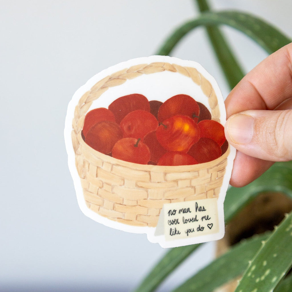 Sticker of a basket with red apples held by a hand, with a plant in the background.