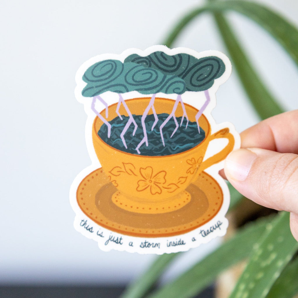 Sticker of a storm in a teacup held by a hand with green leaves in the background