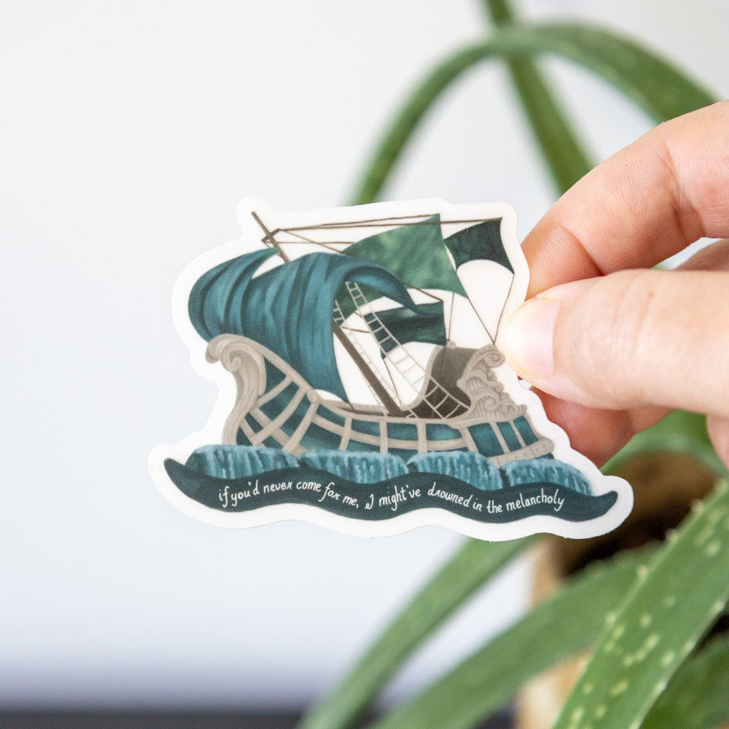 Hand holding a vinyl sticker with a design of a ship on a wave, surrounded by green leaves.
