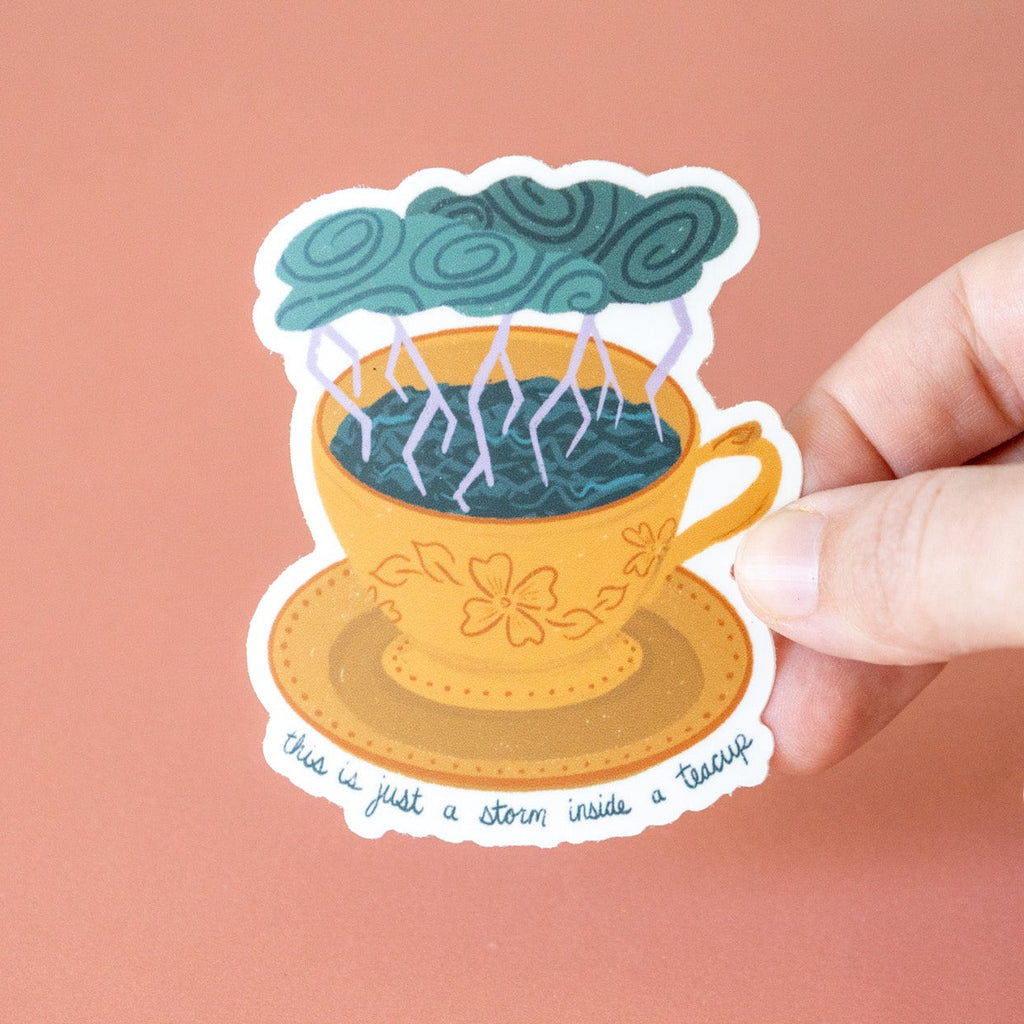 Sticker of a teacup with a storm and text held by a hand on a pink background