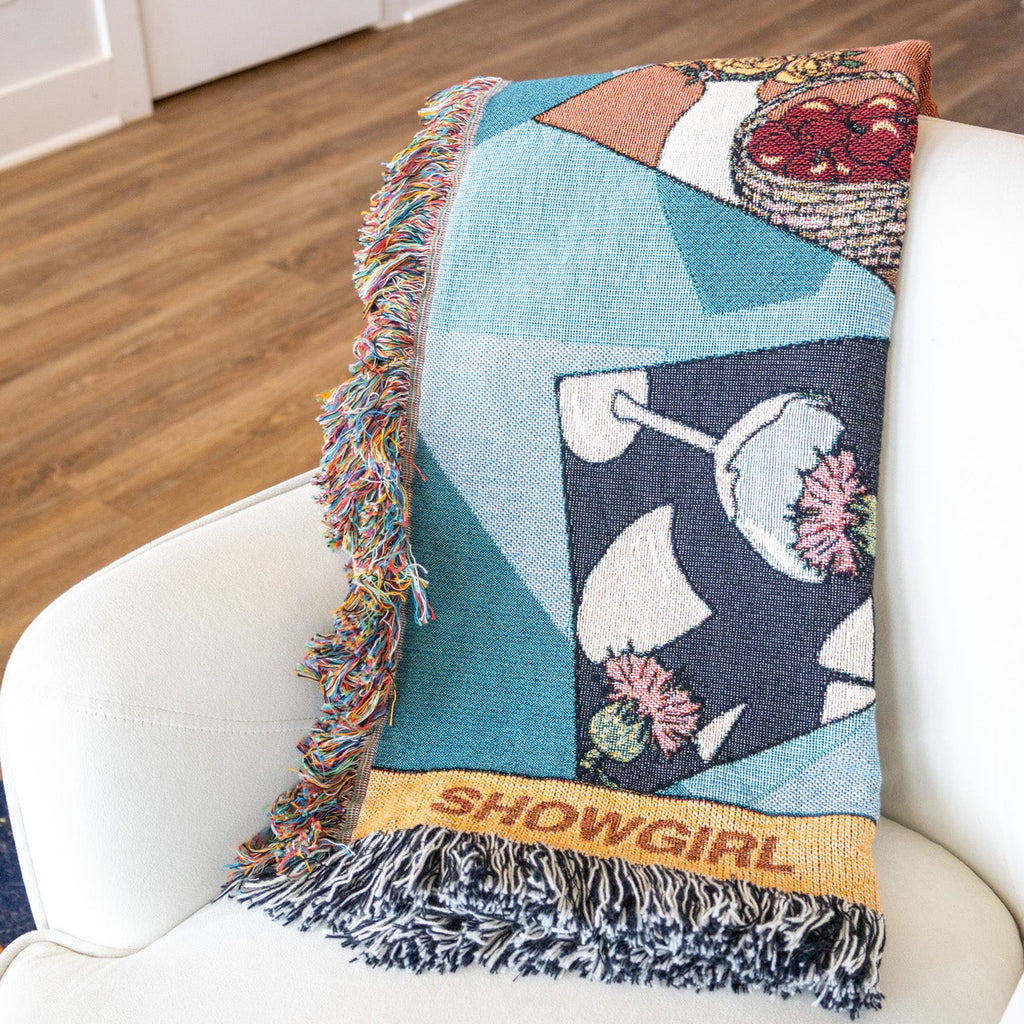 Colorful patterned blanket with 'Showgirl' text draped over a white chair in a room.