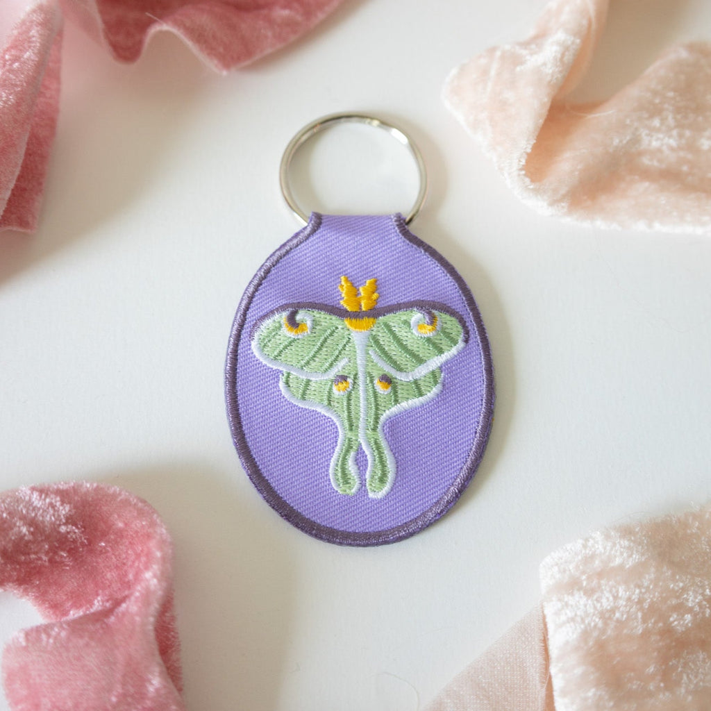 Purple keychain with a green luna moth design on a white background with pink and beige fabric.