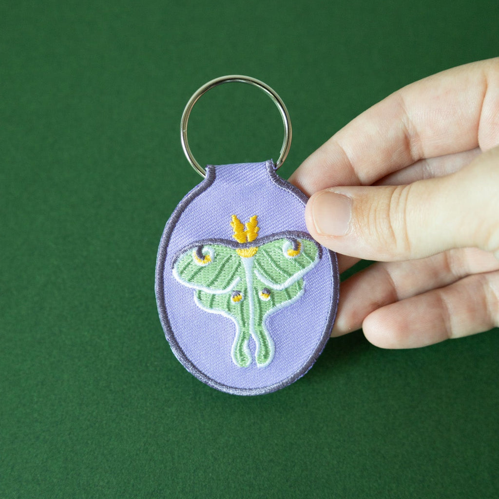 Purple keychain with embroidered luna moth design held by a hand against a green background.