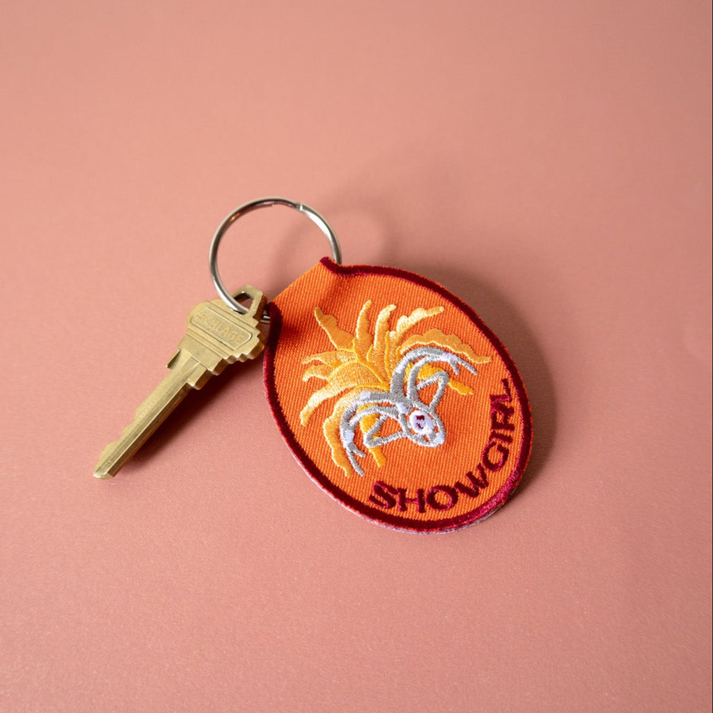 Orange keychain with embroidered design and 'SHOWGIRL' text on a pink background