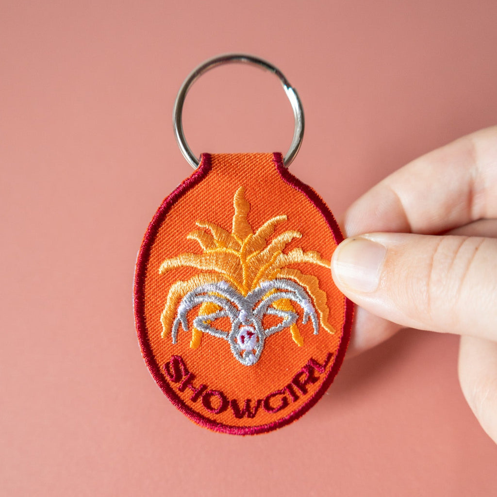 Hand holding an orange keychain with 'Showgirl' design on a pink background