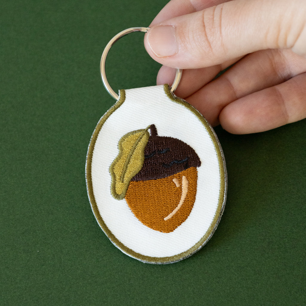 A hand holding a white twill keychain with an acorn embroidered design, displayed on a green background.