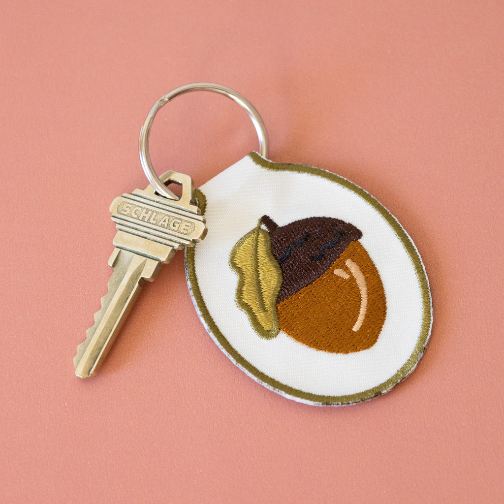 A white twill keychain with an acorn embroidered design, displayed on a pink background.