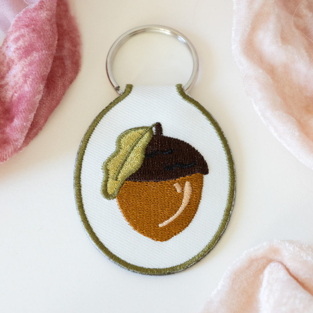 A white twill keychain with an acorn embroidered design, displayed on a white background with pink ribbons.