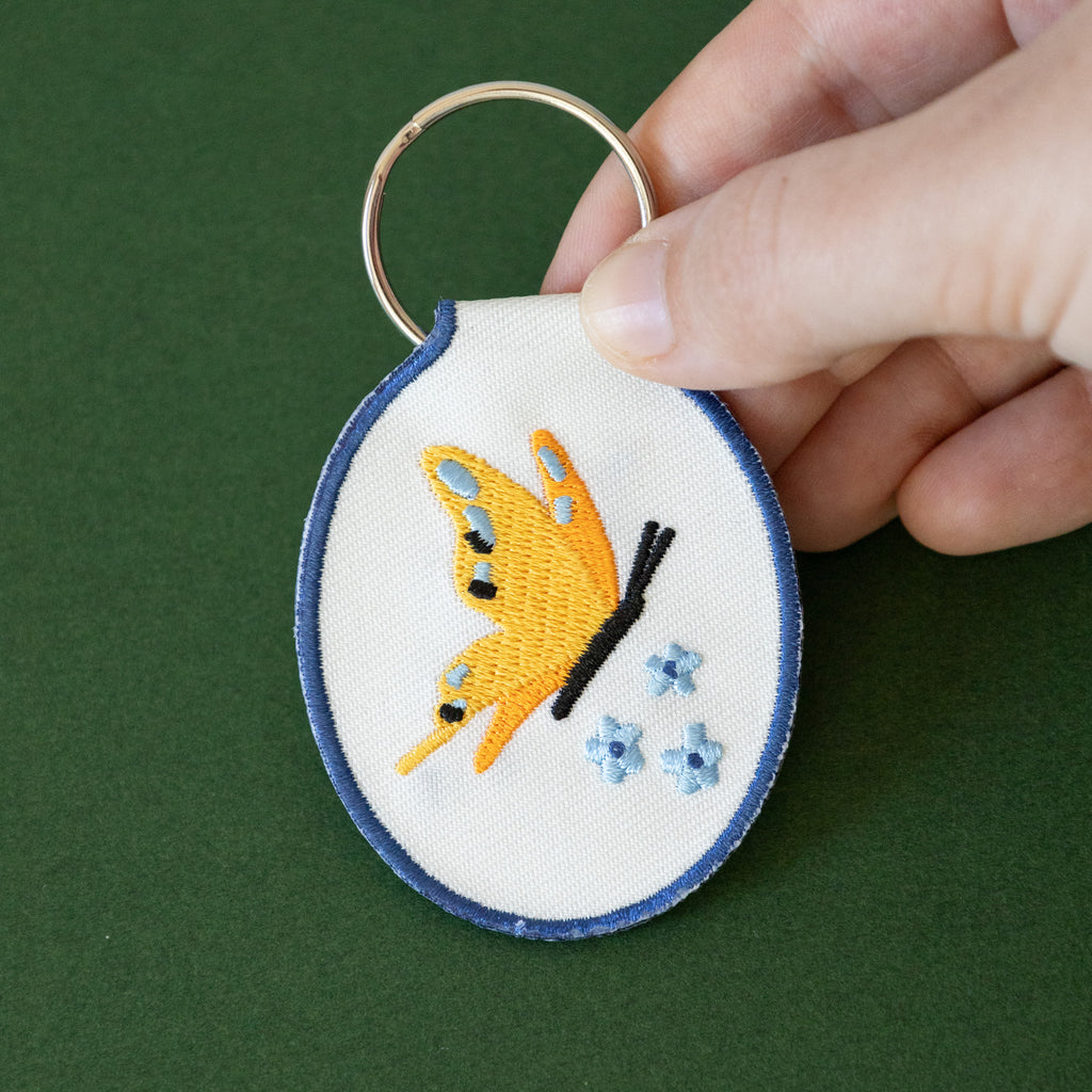 A hand holding a white twill keychain with an embroidered yellow butterfly and blue flowers, displayed on a green background.