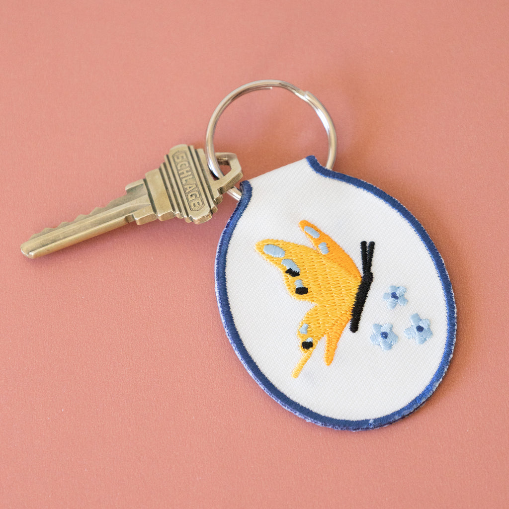 A white twill keychain with an embroidered yellow butterfly and blue flowers, displayed on a pink background.