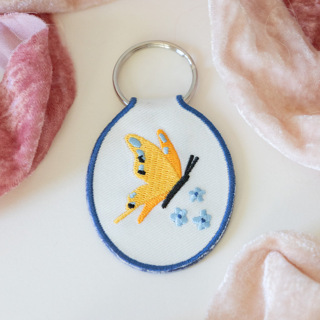 A white twill keychain with an embroidered yellow butterfly and blue flowers, displayed on a white background with pink ribbons.