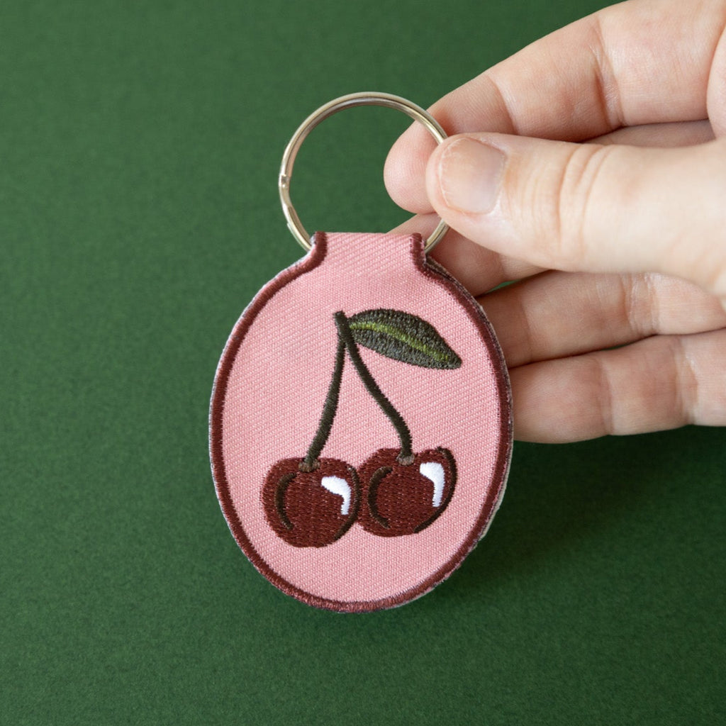 A hand holding a keychain with a cherry embroidery design on a pink cotton twill patch, attached to a metal split key ring, displayed on a green background.