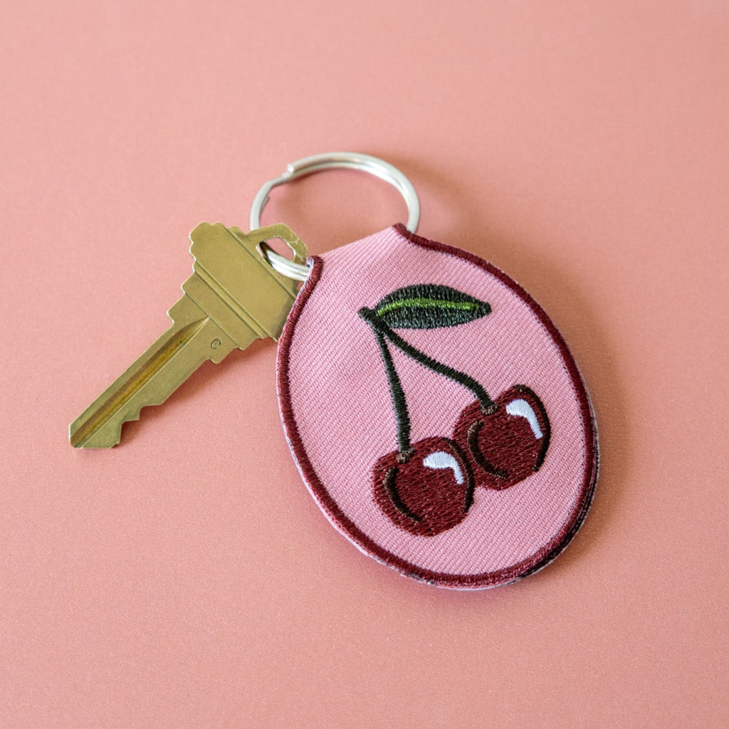 A keychain with a cherry embroidery design on a pink cotton twill patch, attached to a metal split key ring, displayed on a pink background.