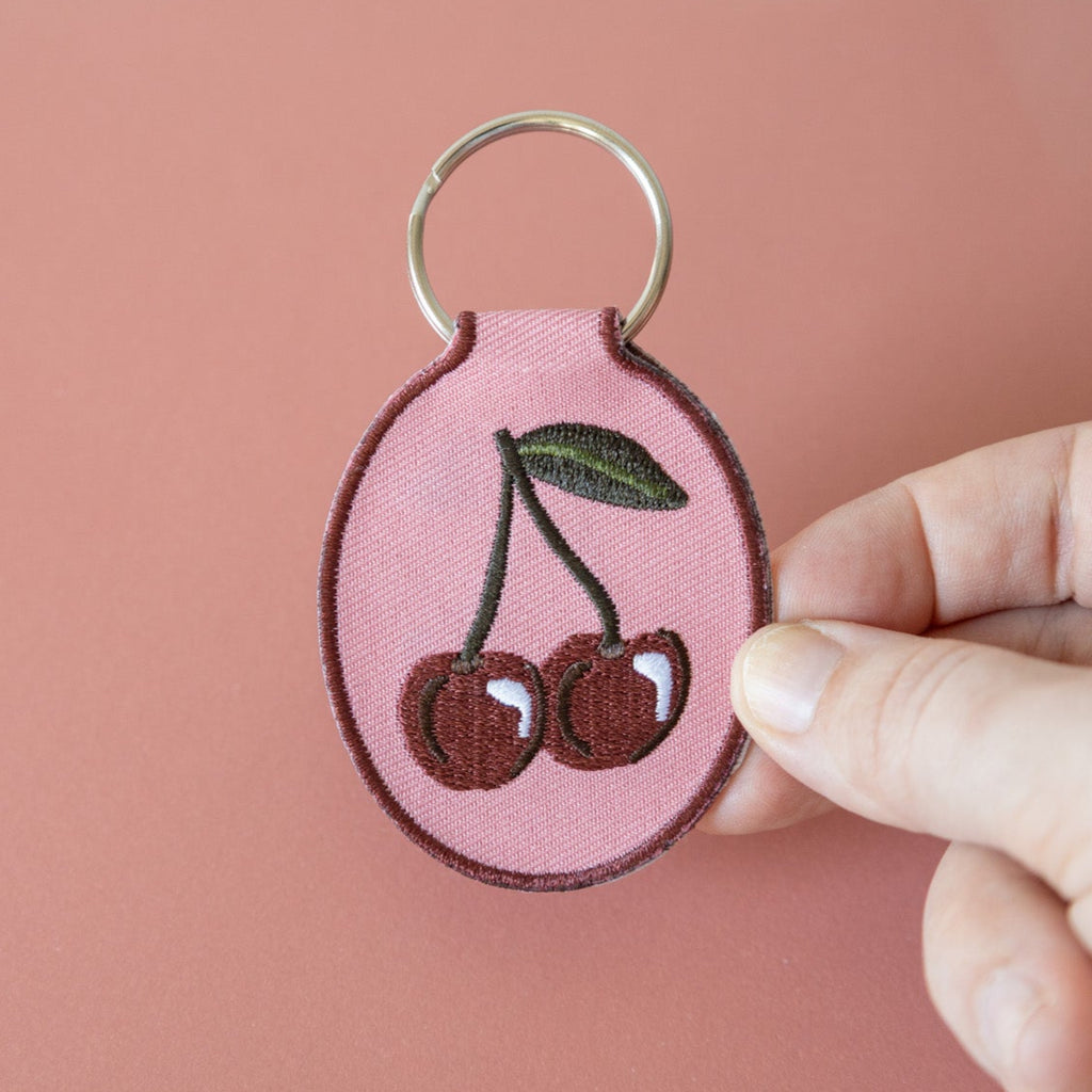 A hand holding a keychain with a cherry embroidery design on a pink cotton twill patch, attached to a metal split key ring, displayed on a pink background.