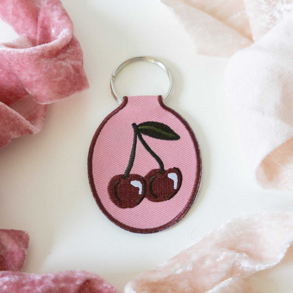 A keychain with a cherry embroidery design on a pink cotton twill patch, attached to a metal split key ring, displayed on a white background with ribbons.