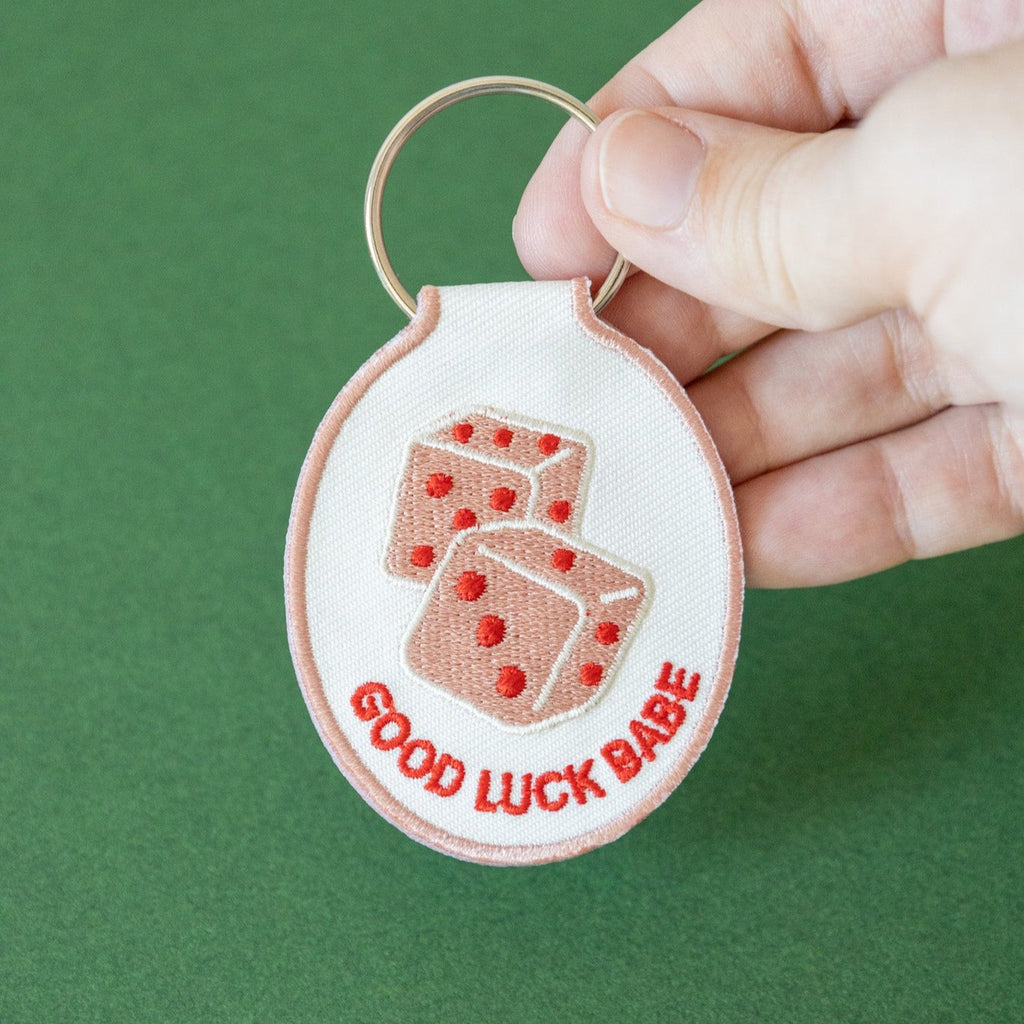 A hand holding white cotton twill keychain with red and pink embroidered dice and the text 'GOOD LUCK BABE' displayed on a green background.
