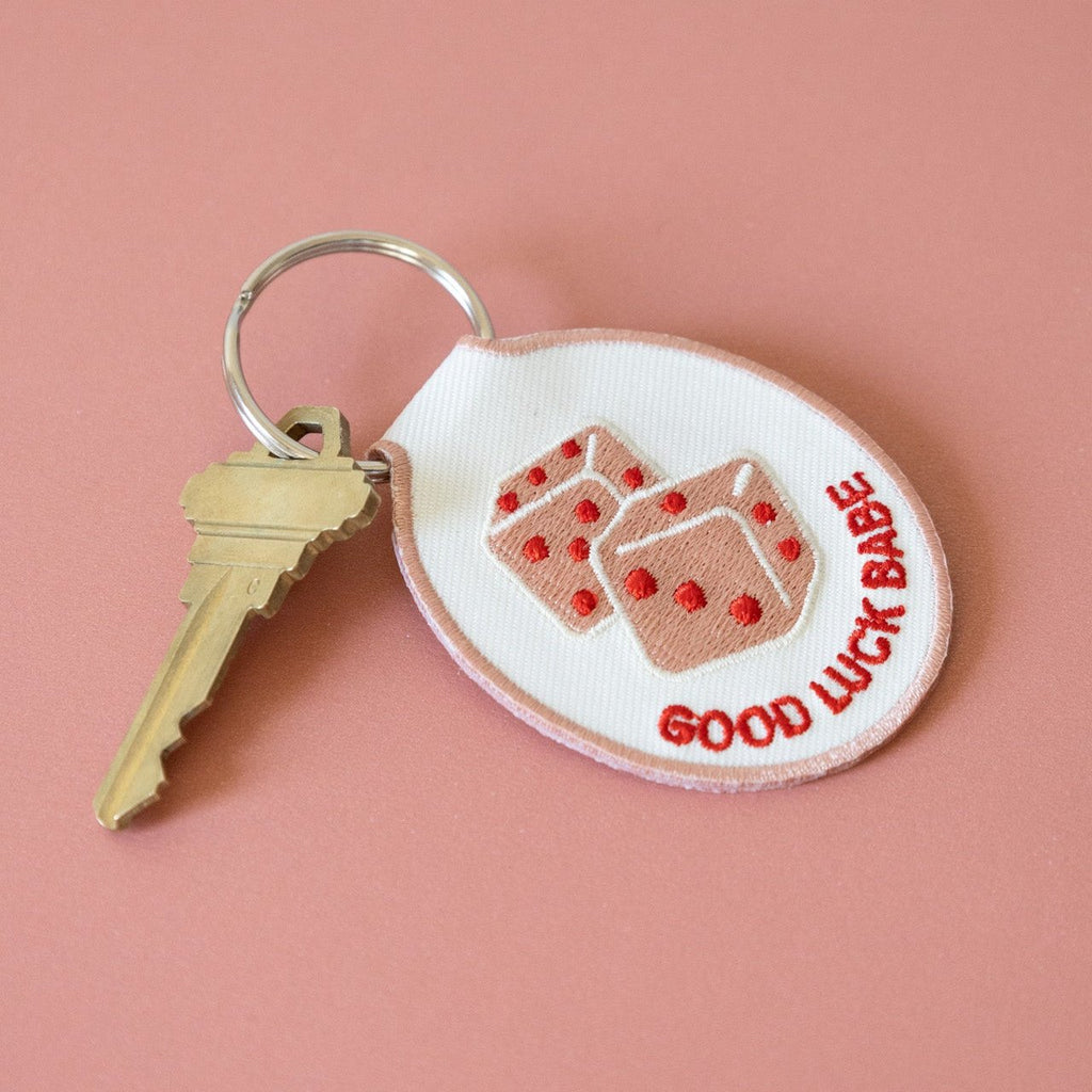 A white cotton twill keychain with red and pink embroidered dice and the text 'GOOD LUCK BABE' displayed on a pink background.
