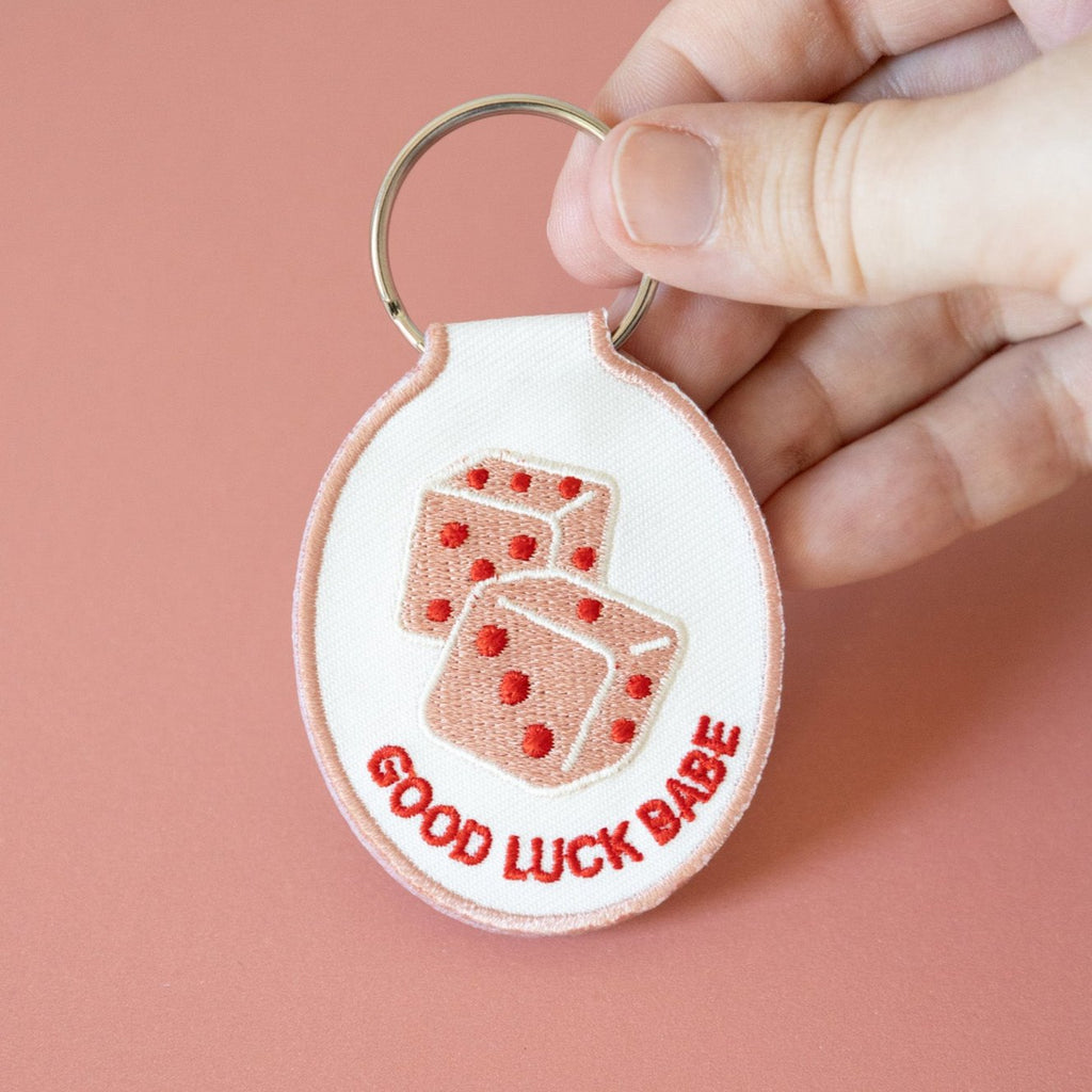 A hand holding white cotton twill keychain with red and pink embroidered dice and the text 'GOOD LUCK BABE' displayed on a pink background.