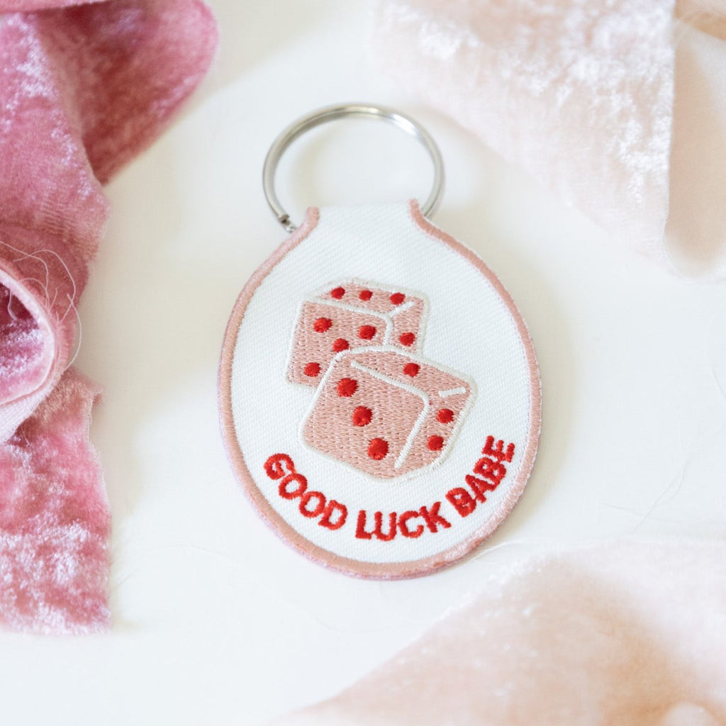 A white cotton twill keychain with red and pink embroidered dice and the text 'GOOD LUCK BABE' displayed on a white background with pink ribbons.