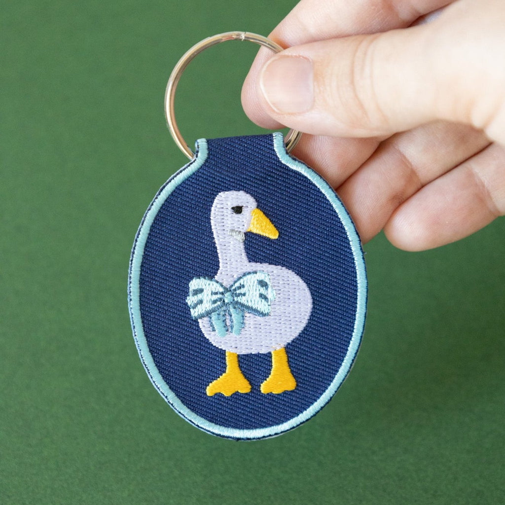 A hand holding a navy twill keychain with an embroidered duck/goose design, displayed on a green background.