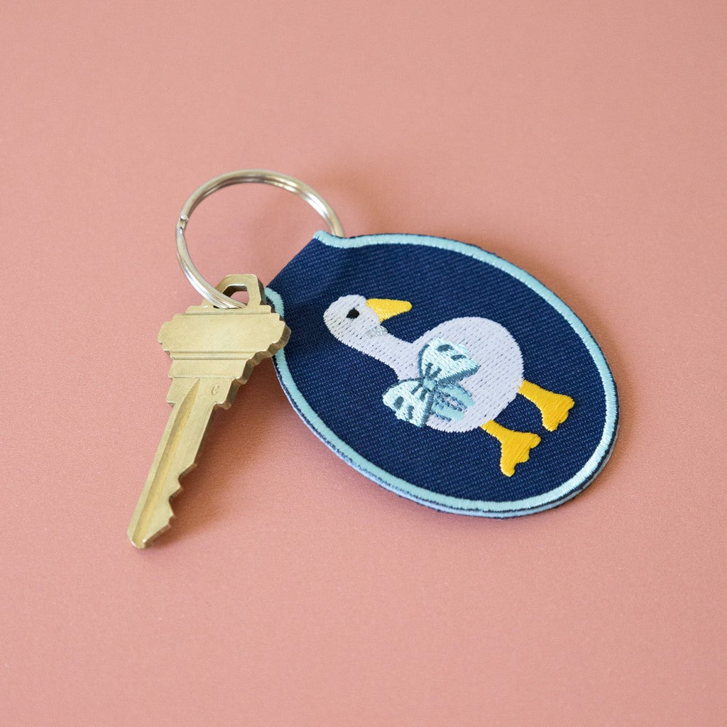 A navy twill keychain with an embroidered duck/goose design, displayed on a pink background.