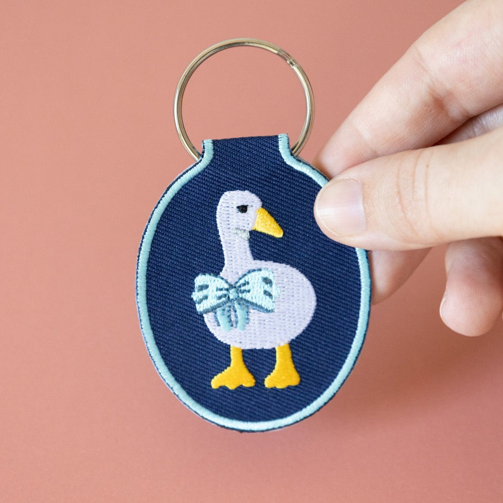 A hand holding a navy twill keychain with an embroidered duck/goose design, displayed on a pink background.
