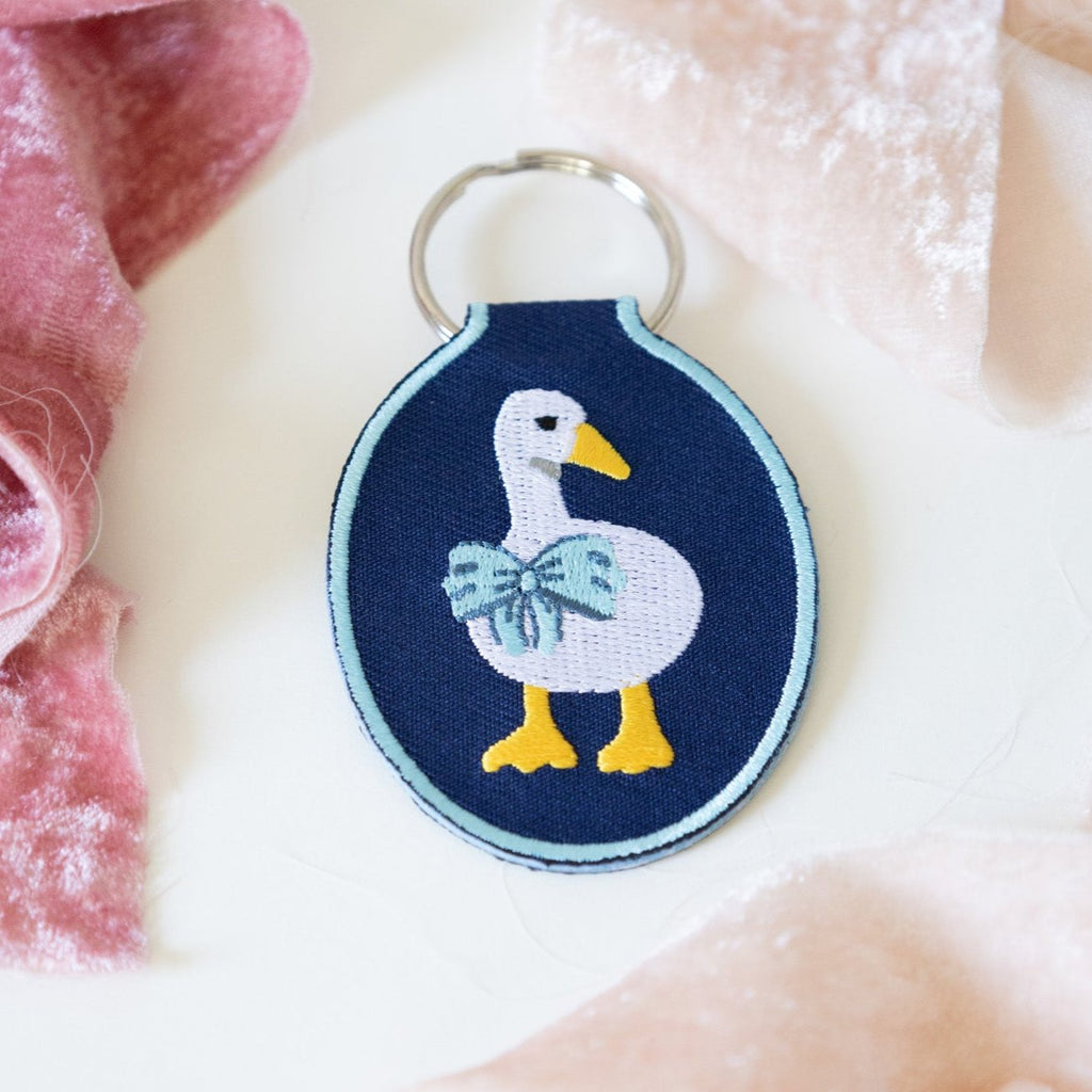 A navy twill keychain with an embroidered duck/goose design, displayed on a white background with pink ribbons.