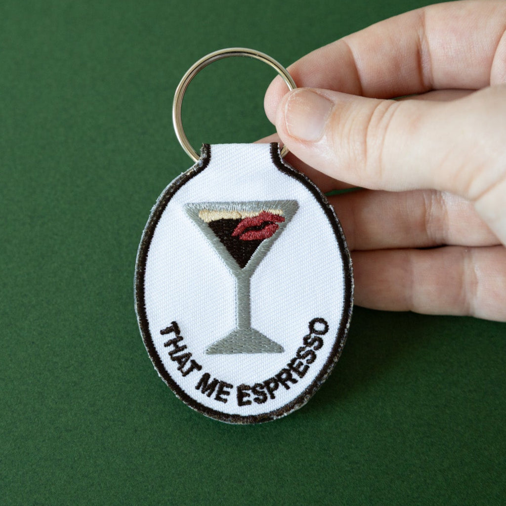 A hand holding an embroidered keychain with a design of an espresso martini with a kiss and the phrase 'that me espresso' on it, attached to a metal key ring, displayed on a green background.