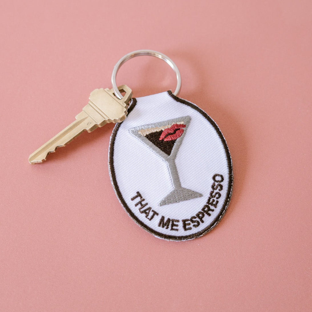 An embroidered keychain with a design of an espresso martini with a kiss and the phrase 'that me espresso' on it, attached to a metal key ring, displayed on a pink background.