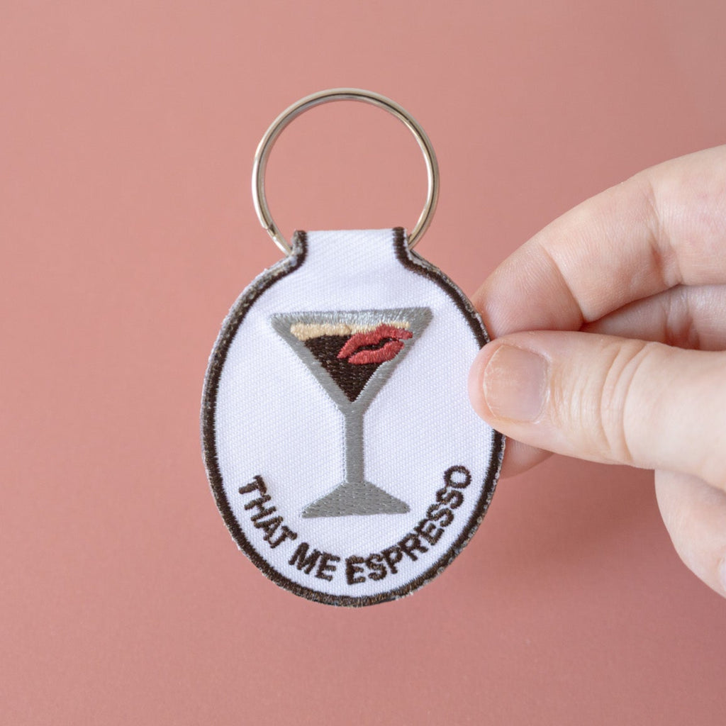 A hand holding an embroidered keychain with a design of an espresso martini with a kiss and the phrase 'that me espresso' on it, attached to a metal key ring, displayed on a pink background.