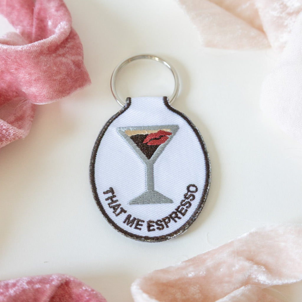 An embroidered keychain with a design of an espresso martini with a kiss and the phrase 'that me espresso' on it, attached to a metal key ring, displayed on a white background with pink ribbons.