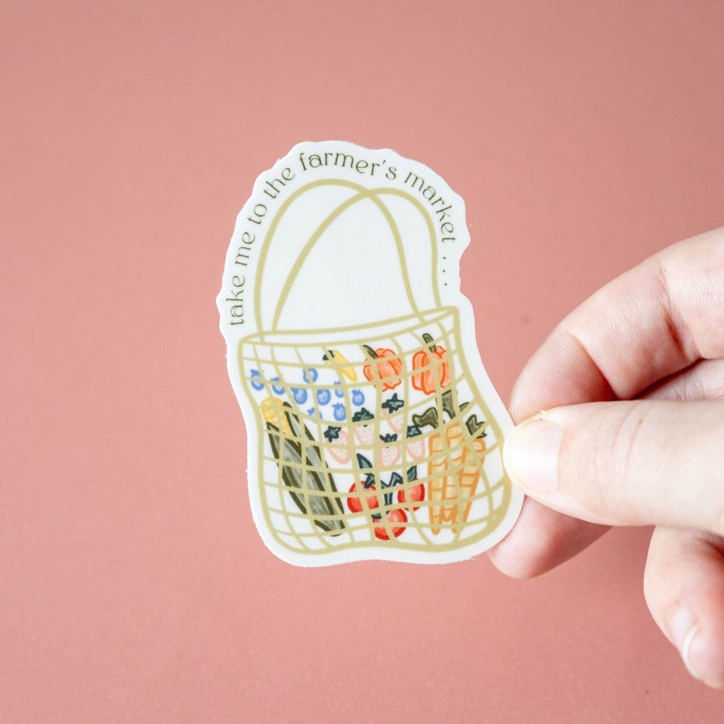 Hand holding a sticker of a basket with fresh produce against a pink background