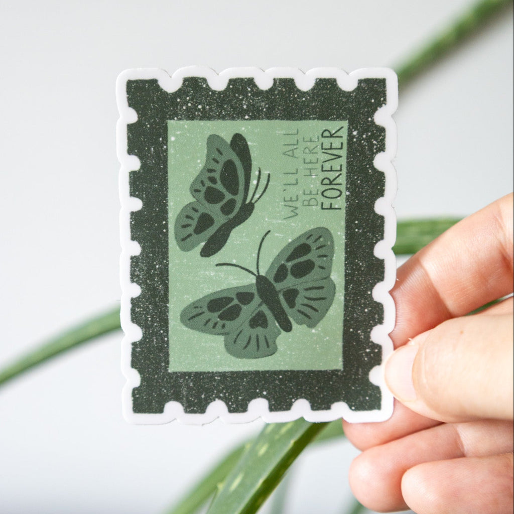 Hand holding a decorative stamp sticker with butterfly design and text that reads "we'll all be here forever" against a white background