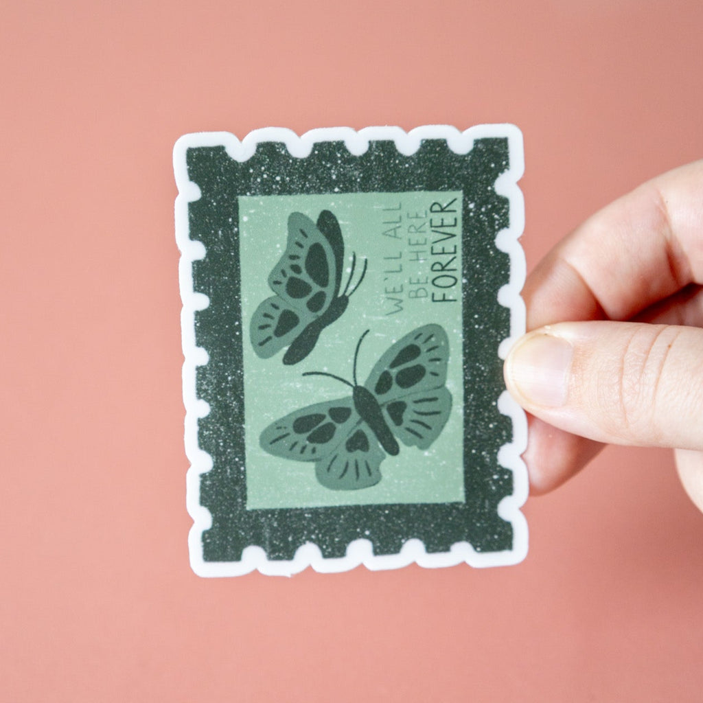 Hand holding a decorative stamp sticker with butterfly design and text that reads "we'll all be here forever" against a pink background