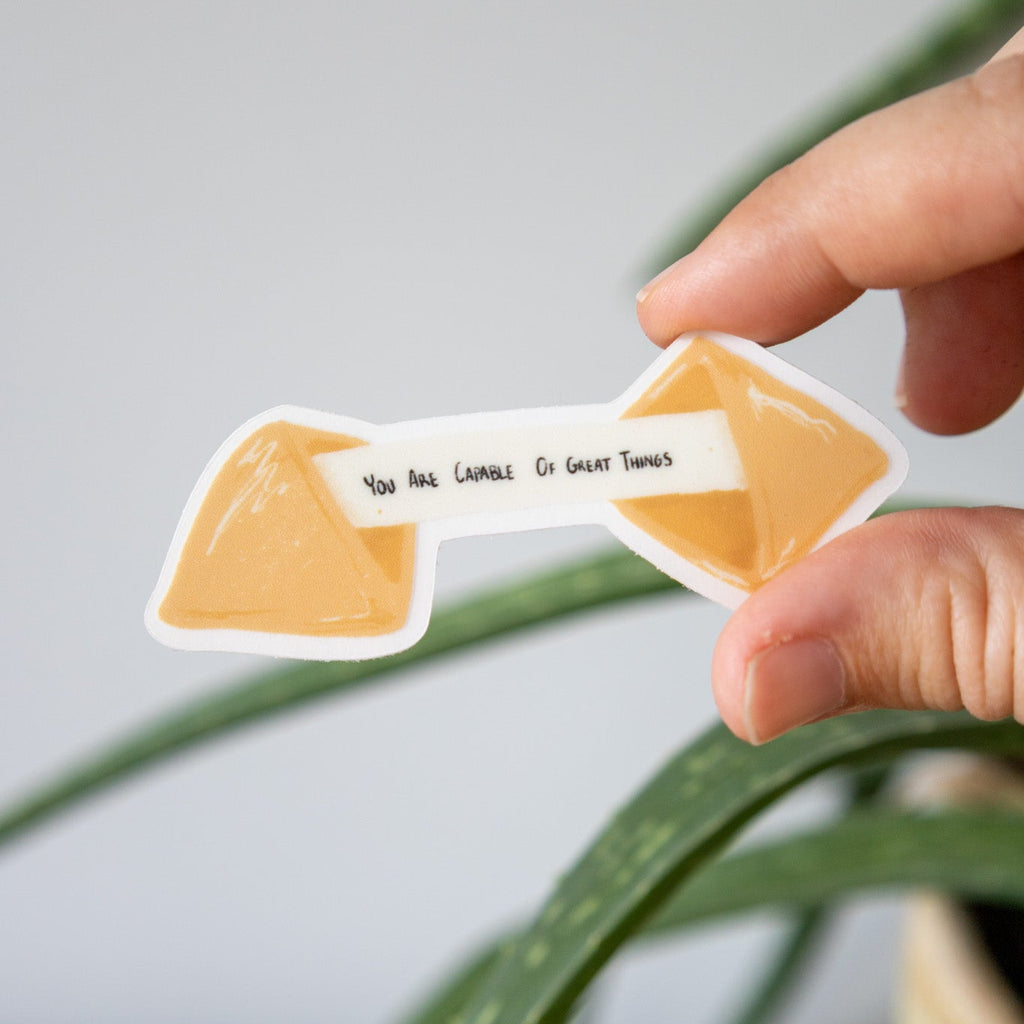 fortune cookie sticker that says "you are capable of great things held by a hand with a plant in the background