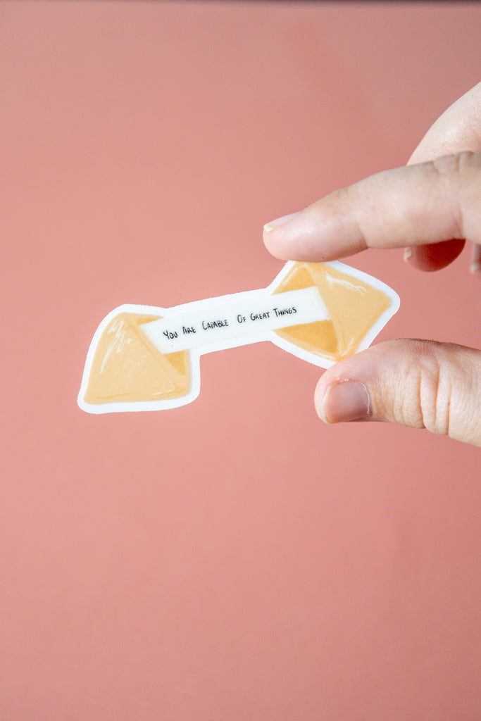 fortune cookie sticker that says "you are capable of great things held by a hand against a pink background