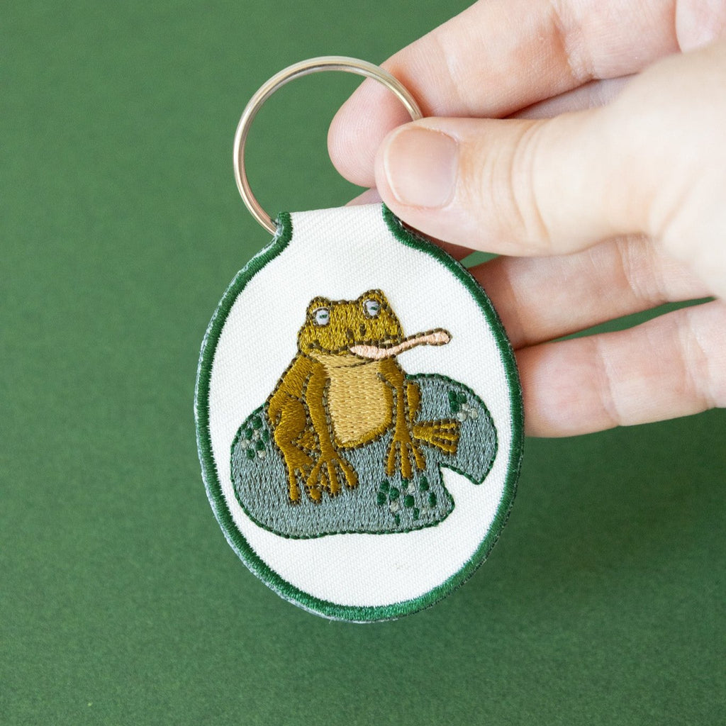 A hand holding a cream twill keychain with an embroidered frog on a lily pad design, displayed against a green background.