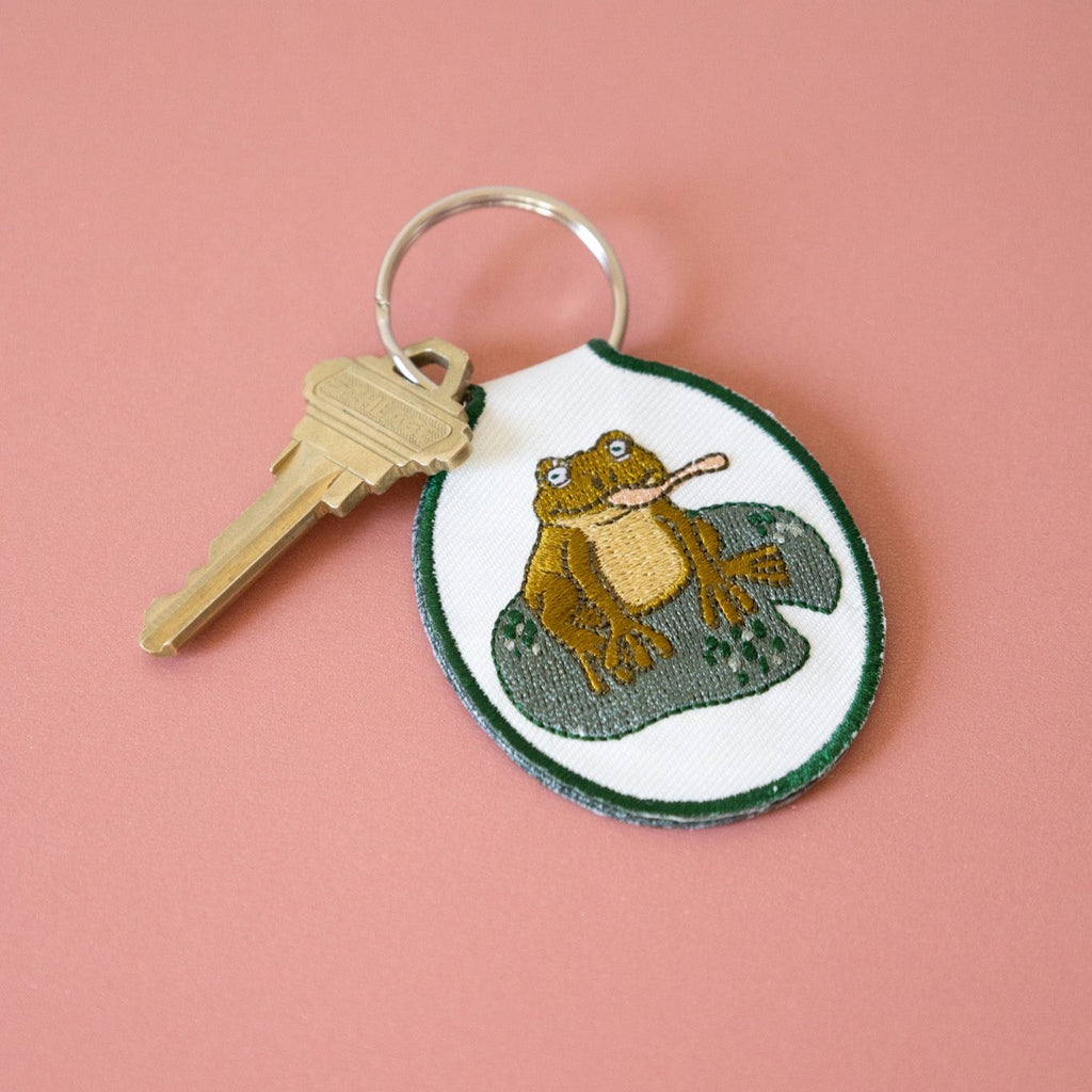 A cream twill keychain with an embroidered frog on a lily pad design, displayed against a pink background.