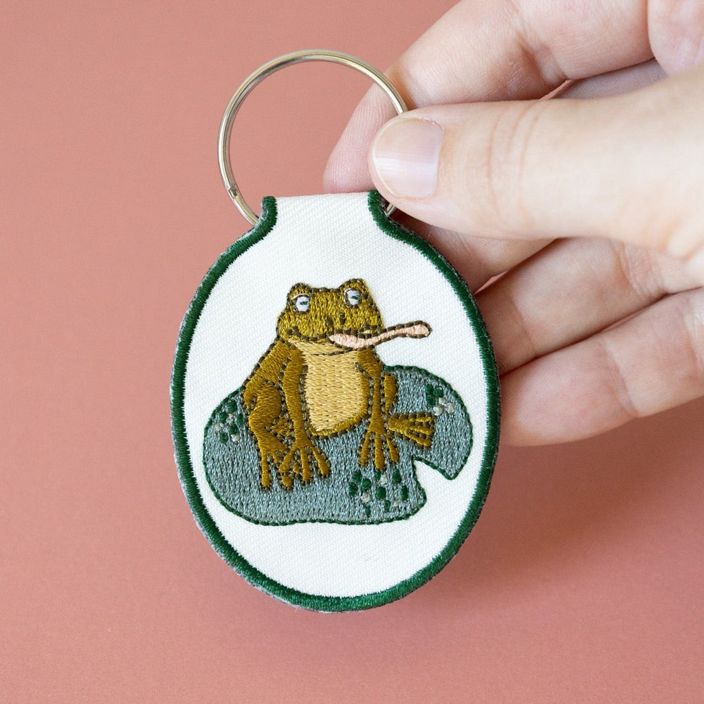 A hand holding a cream twill keychain with an embroidered frog on a lily pad design, displayed against a pink background.
