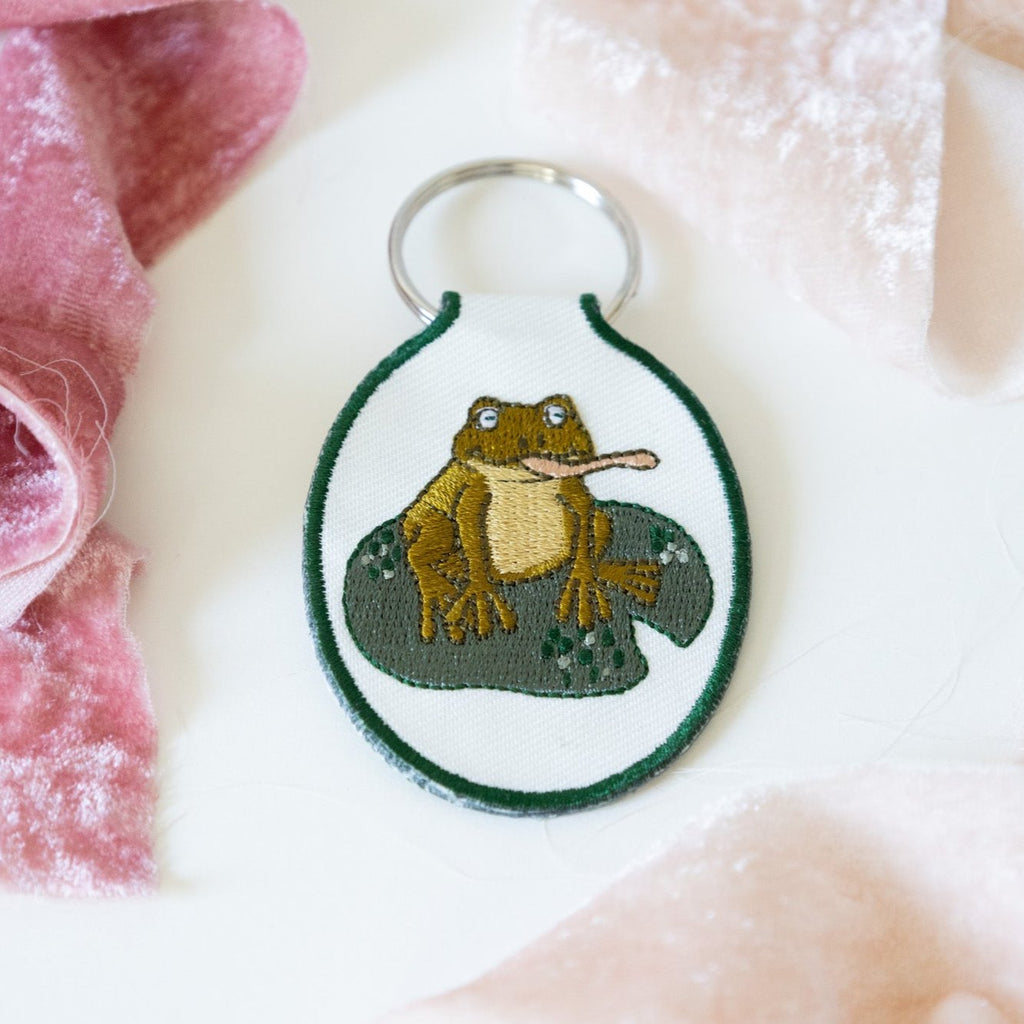 A cream twill keychain with an embroidered frog on a lily pad design, displayed against a white background with pink ribbons.