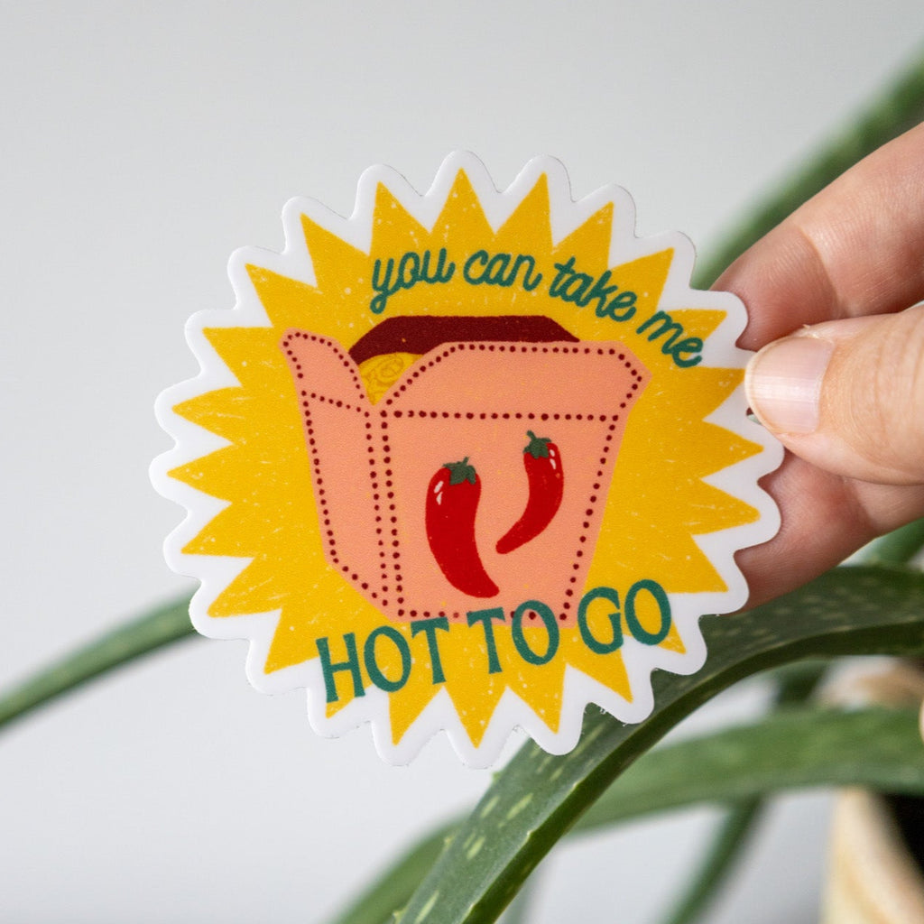 Sticker with a design of a take out box and red peppers, text 'you can take me HOT TO GO', held by a hand against a plant background.