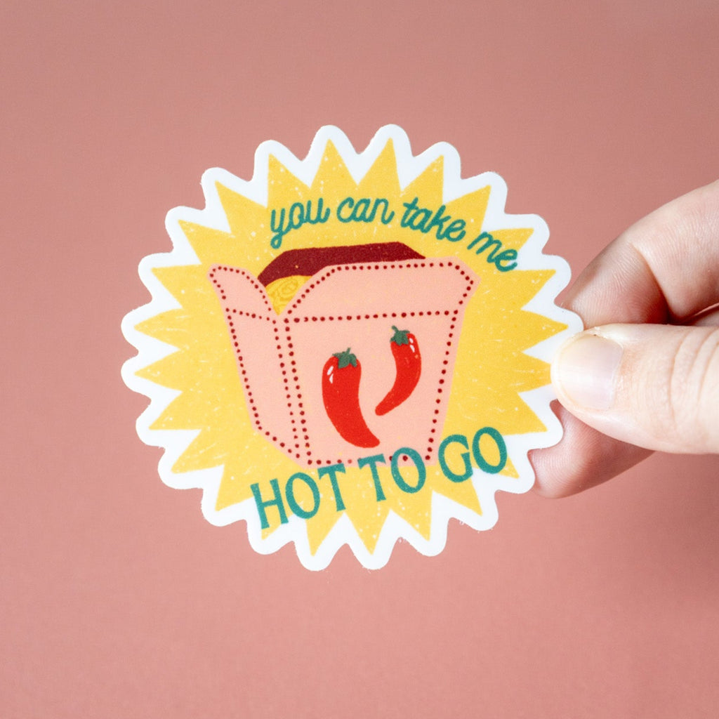 Sticker with a design of a take out box and red peppers, text 'you can take me HOT TO GO', held by a hand against a pink background.