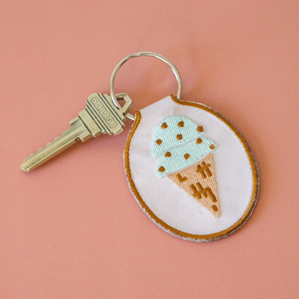 A light pink keychain with a mint chocolate chip ice cream cone embroidered design, displayed on a pink background.