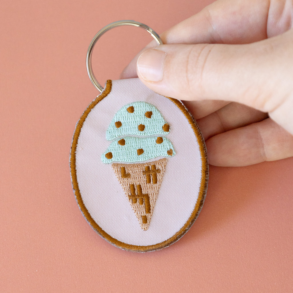 A hand holding a light pink keychain with a mint chocolate chip ice cream cone embroidered design, displayed on a pink background.