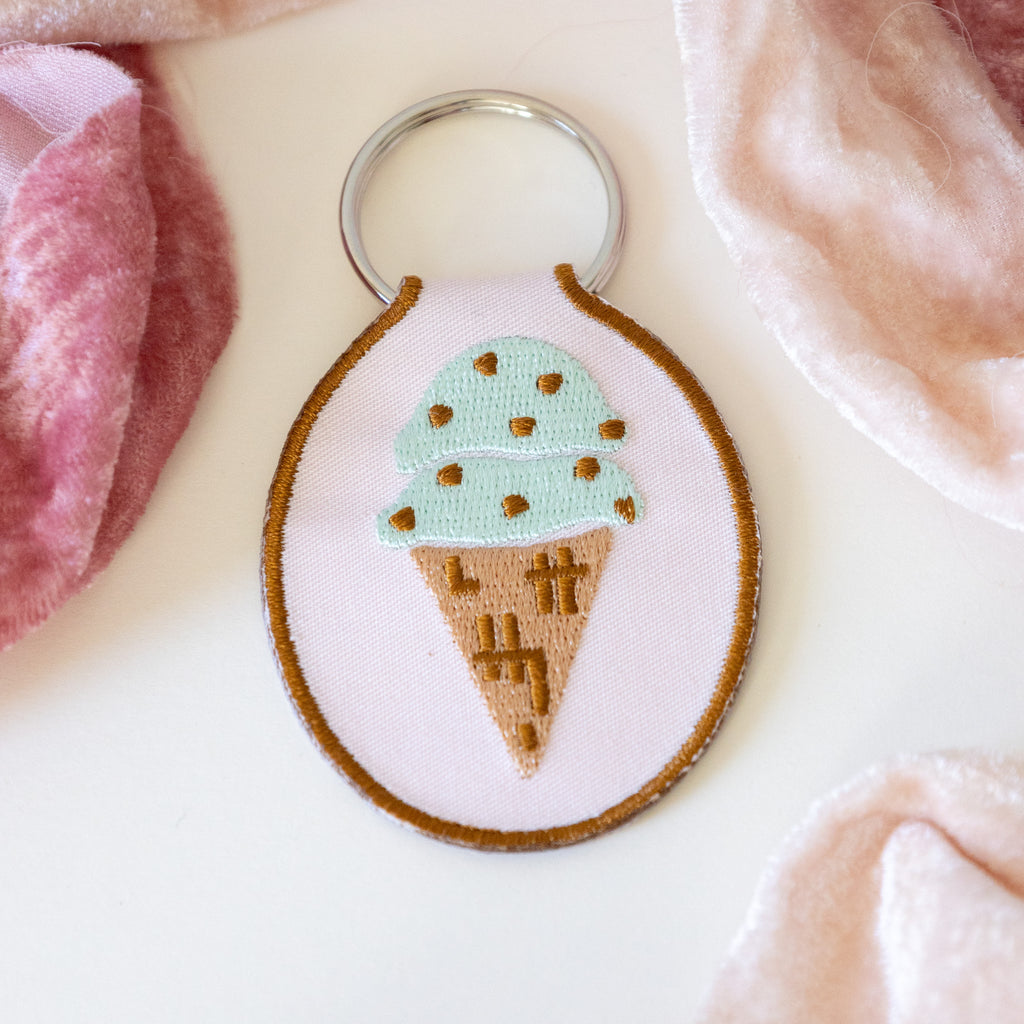 A light pink keychain with a mint chocolate chip ice cream cone embroidered design, displayed on a white background with pink ribbons.