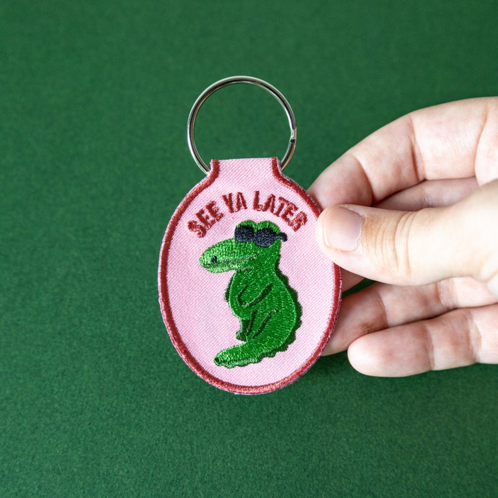 Pink embroidered keychain with green alligator design and text that says "See Ya Later" held by a hand against a green background.