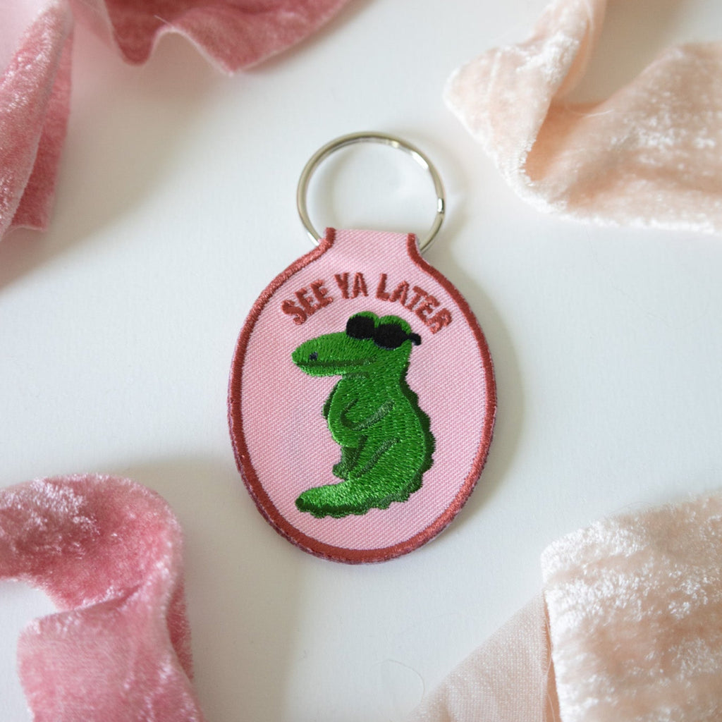 Pink keychain with green alligator and text 'See Ya Later' on a white background with pink ribbons.