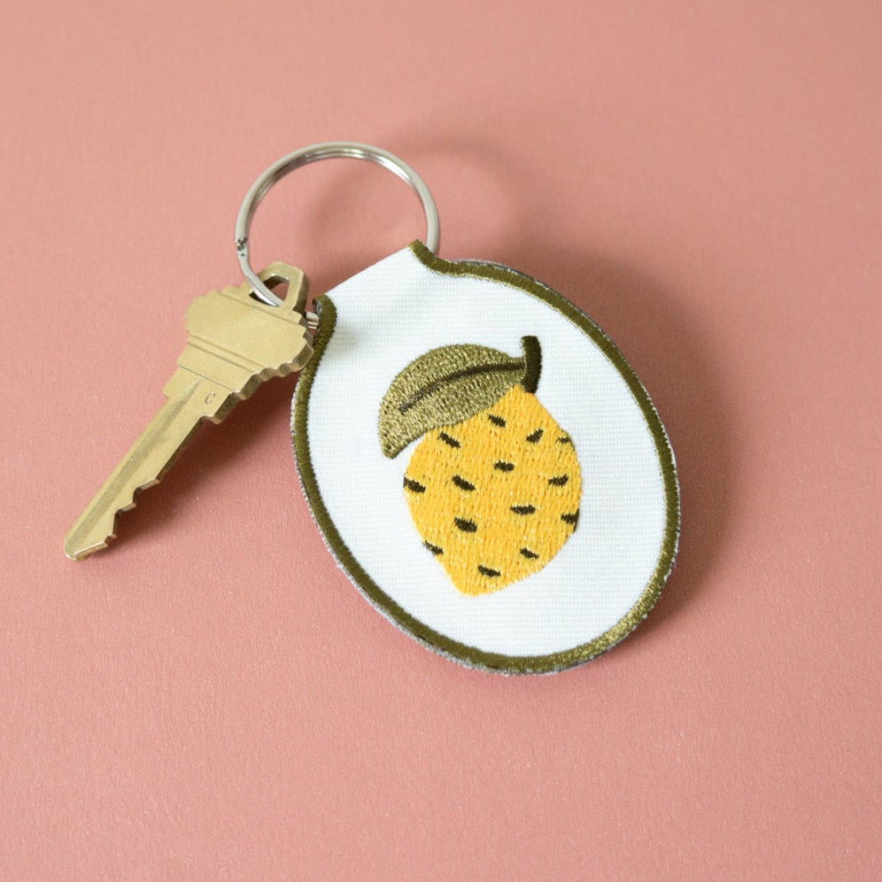 Embroidered keychain with lemon design on a cream twill fabric against a pink background.